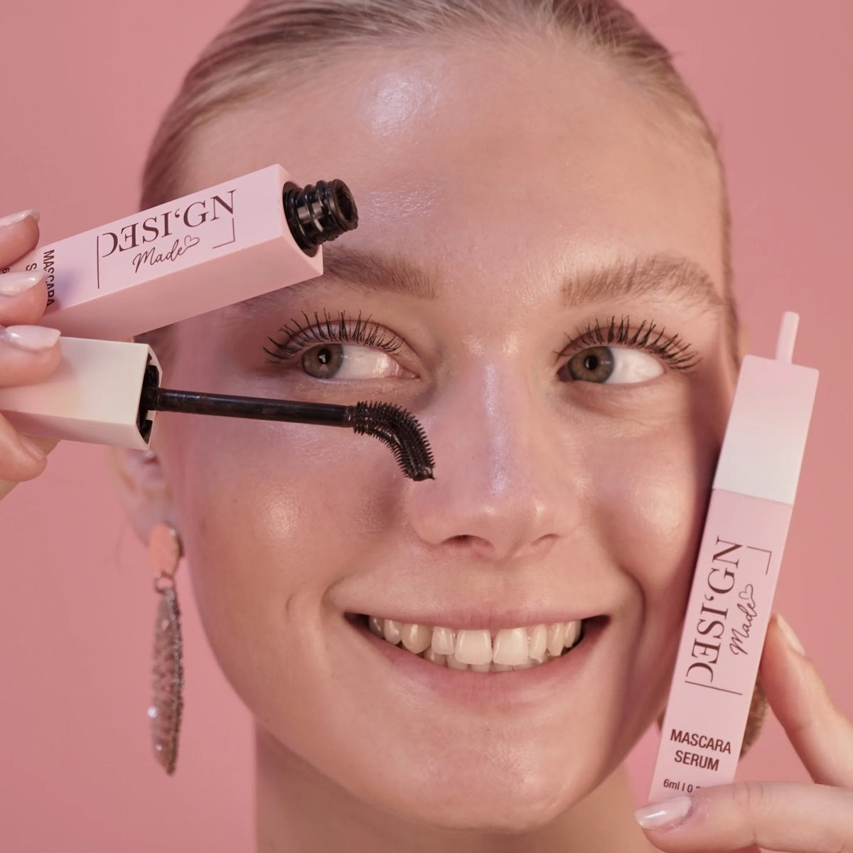 Frau hält zwei roséfarbene Tuben der TUBING MASCARA mit Wachstumsserum von DESI'GN MADE neben ihr Gesicht, eine davon geöffnet mit der Bürste
